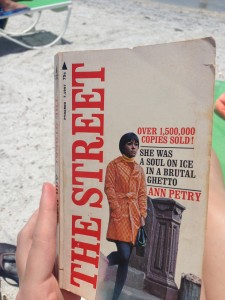 A hand holds a paperback copy of Ann Petry's The Street in the forefront of the image, with sand and beach chairs in the background. The tagline on the cover reads, "She was a soul on ice in a brutal ghetto" and "over 1,500,000 copies sold!"