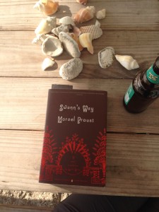 A paperback copy of Swann's Way (brown cover, with red ornate design details), on a wooden table with several sea shells and a bottle of beer. 