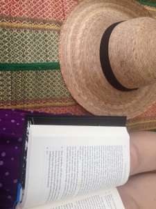 An open book on the lap of someone sitting on a colorful woven mat, beside a round straw hat. 