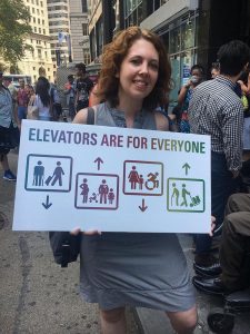 Photo of Jessica Murray holding a sign that says "Elevators are for Everyone" with icons of various people who use elevators inside boxes with arrows representing elevators moving up and down. The icons represent a person with crutches, a person rolling a suitcase, a pregnant woman, a person with a heart condition, a young girl, a woman and young boy, a wheelchair user, a person with a cane, and a delivery person rolling a cart with packages. 