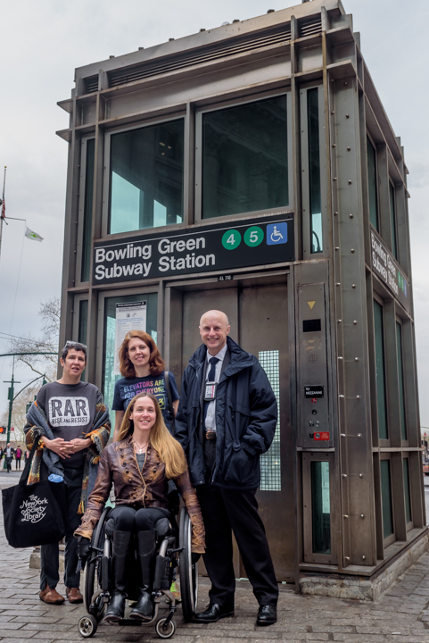 Photo of four people in front of the Bowling Green subway station elevator, one of few wheelchair accessible stations in downtown Manhattan. Front, April Coughlin, who uses a manual wheelchair. Back, Jennifer Marie Bartlett, Jessica Murray, and Andy Byford, NYCT President.