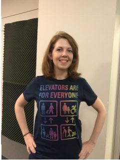 Jessica Murray wearing a navy blue t-shirt with a rainbow-colored graphic that says Elevators are for Everyone, and has icons of a pregnant woman with a stroller, young children, people with crutches, an older person with a cane, people with invisible disabilities, and able-bodied people rolling luggage and carts.