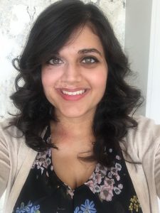 A headshot of Ashanka Kumari. In the picture, she is wearing a beige cardigan and a black top with flowers on it, and she is smiling in front of a white background.