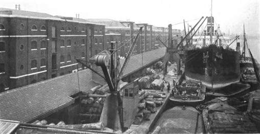 Black and white photograph of a dock with a tall ship with cargo being loaded or unloaded beside it, on the dock. Brick buildings flank the dock.